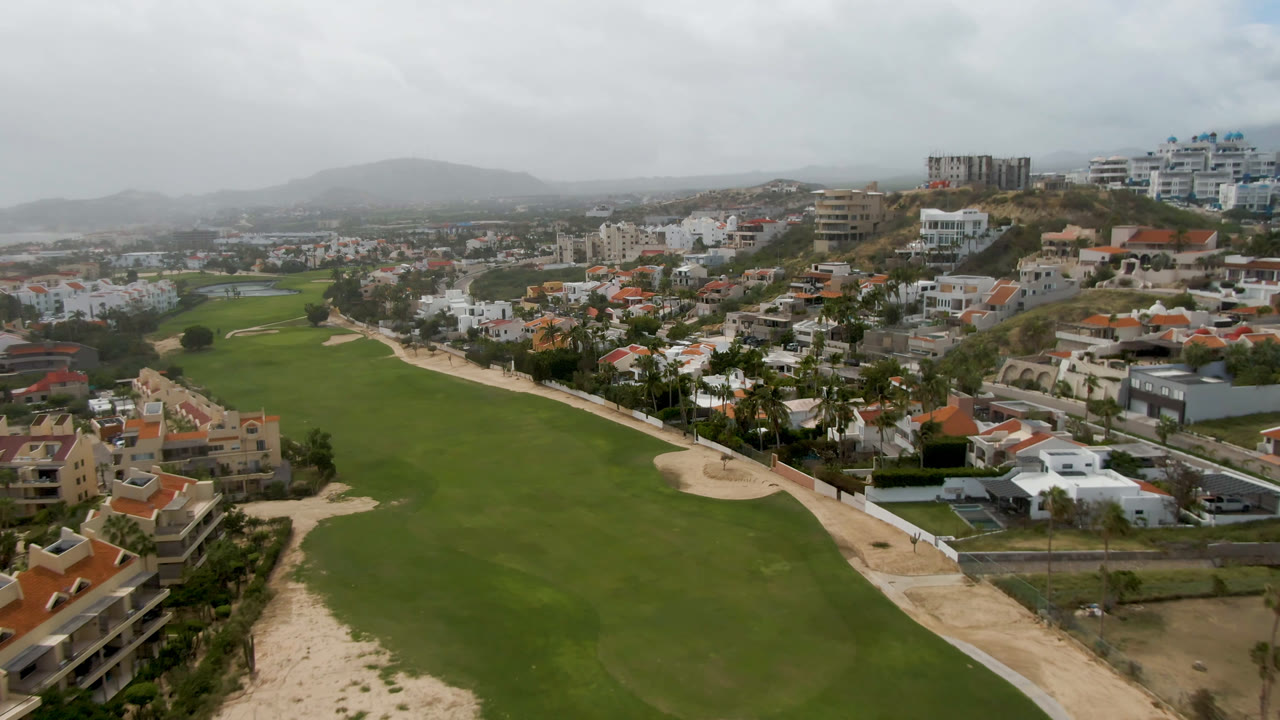 Aerial frame from San Jose del Cabo showing the central fairway, pale paths, surrounding houses, a roadway edge, muted hillside terrain, and no visible people.