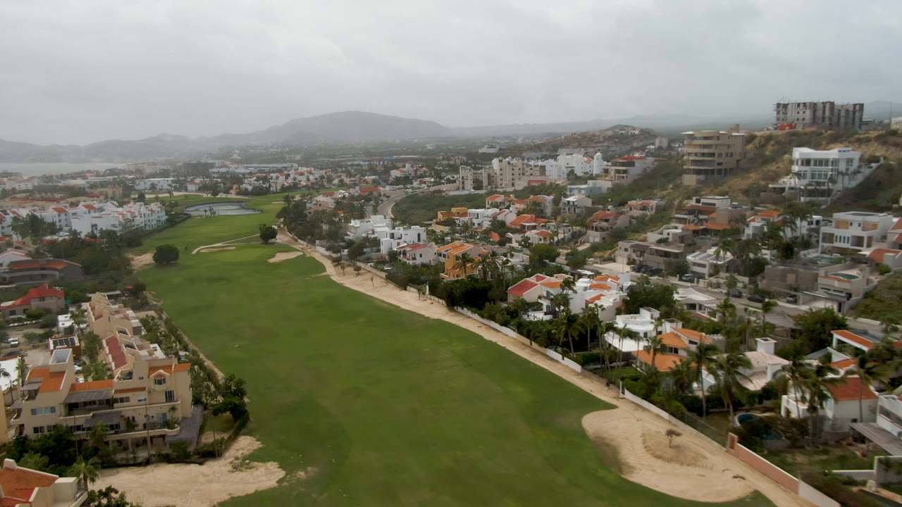 Second aerial frame in San Jose del Cabo showing the fairway running through the foreground, homes along the edge, hillside construction, distant water, and no visible people.