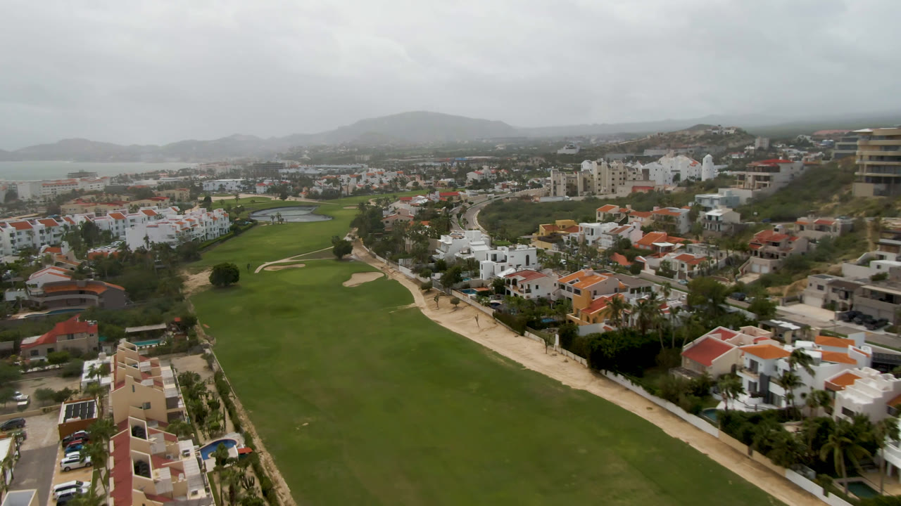 Aerial view of a golf course beside white residential buildings and distant water.