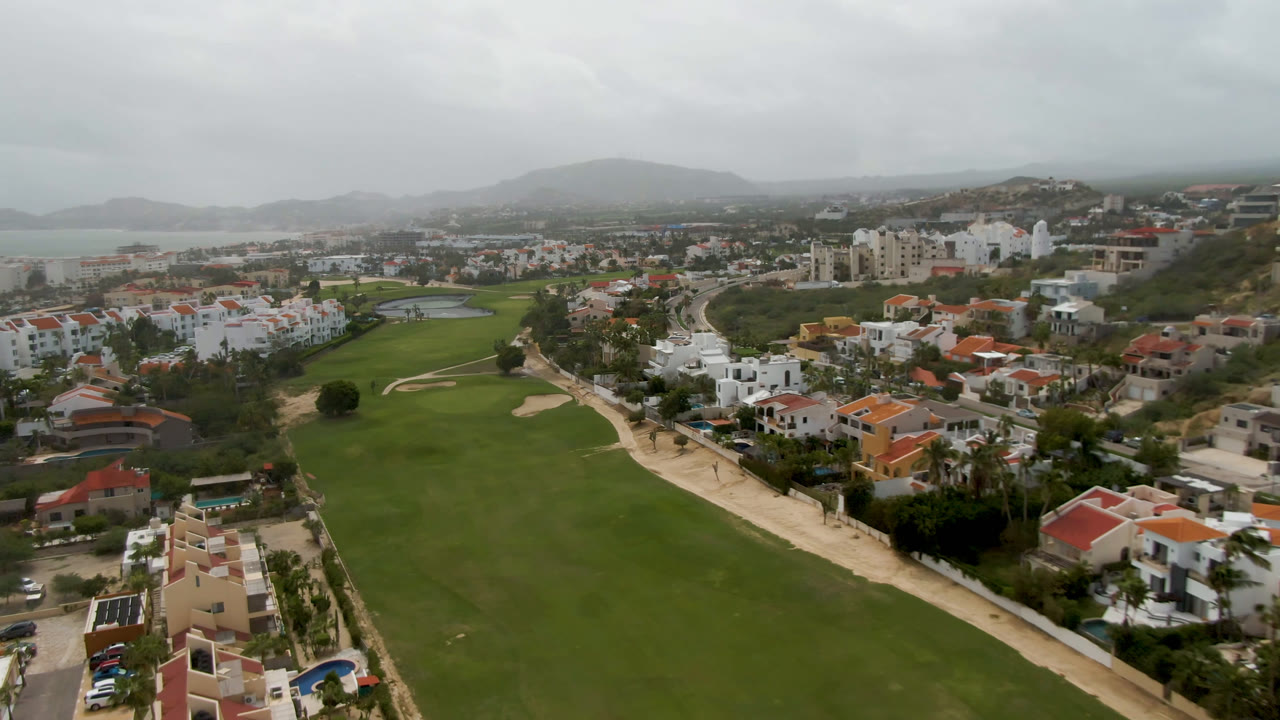 Aerial view in San Jose del Cabo with the fairway stretching toward white buildings, nearby roads, red-roofed houses, hazy hills, distant water, and no visible people.