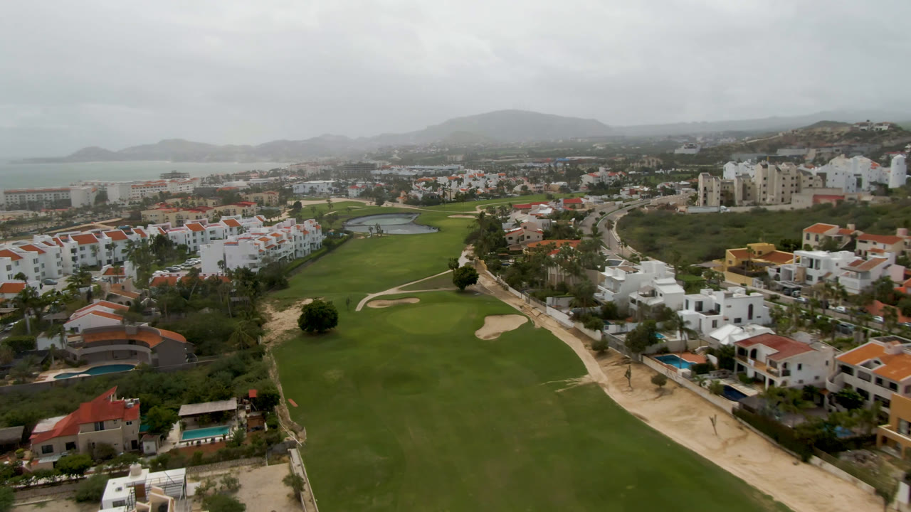 Opening aerial frame over San Jose del Cabo, Mexico, showing a green golf fairway, sand bunkers, white homes, red roofs, hazy hills, coastal water, and no visible people.