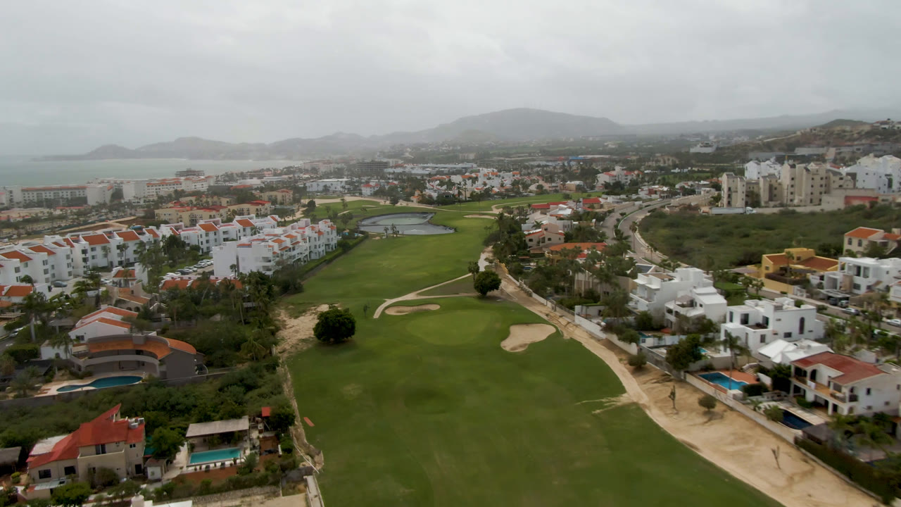 Aerial frame in San Jose del Cabo with the fairway centered, a curved road along the right, white residential buildings, a small lake or water feature, distant hills, and no visible people.