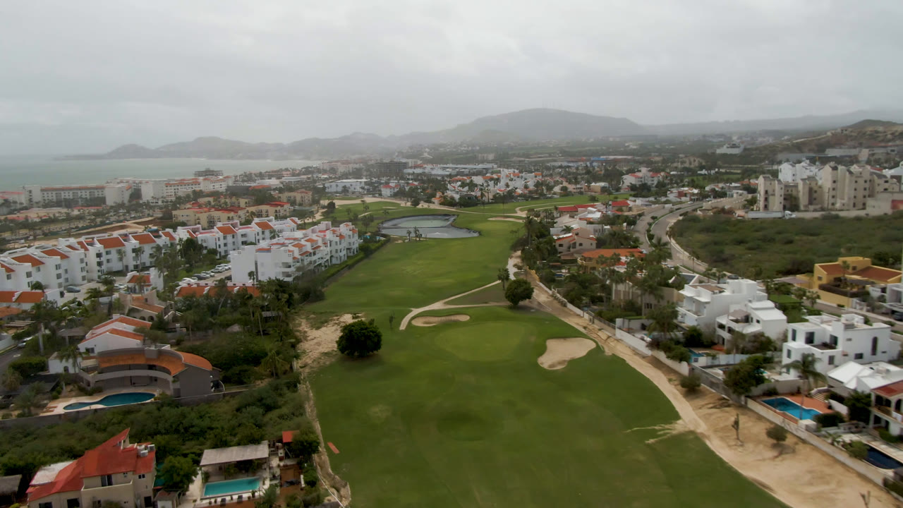 Drone-height view of fairway, bunkers, homes, pools, hills, and coast.