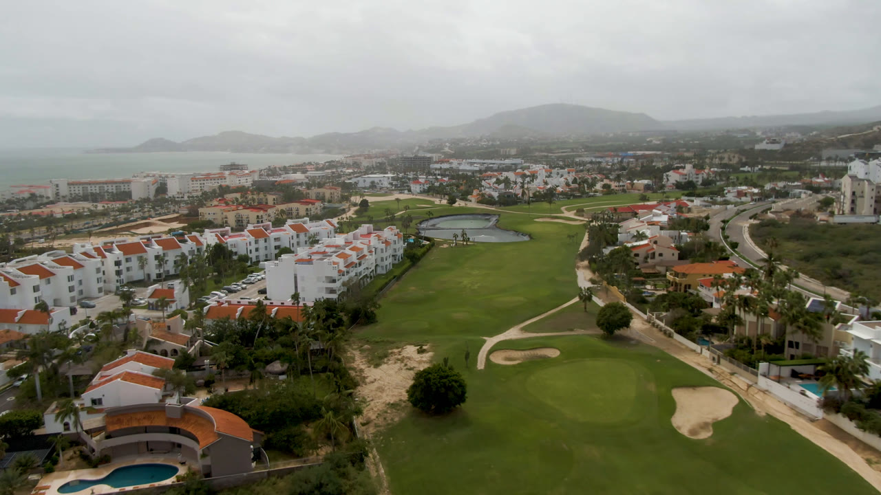 Aerial frame from San Jose del Cabo showing green fairways, sand bunkers, white red-roof buildings, roads, distant water, hazy hills, and no visible people.