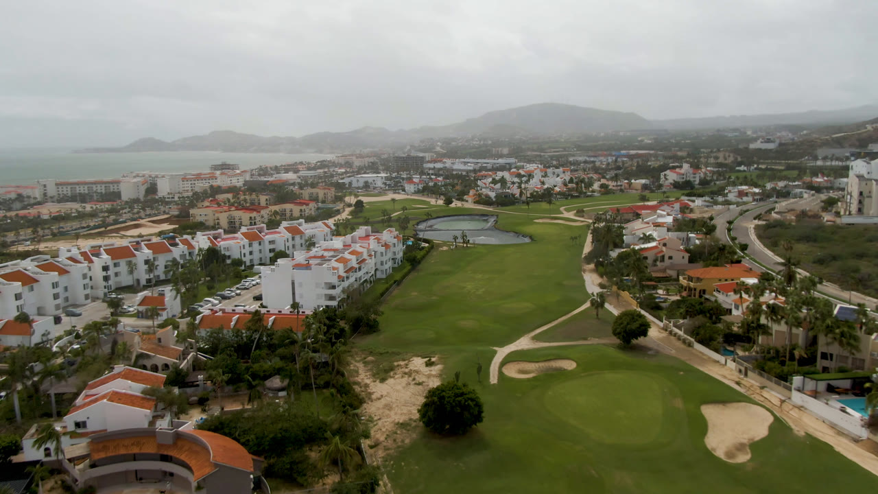 San Jose del Cabo aerial view with a pond near the golf course, rows of white buildings, curving roads, distant hills, gray sky, and no visible people.