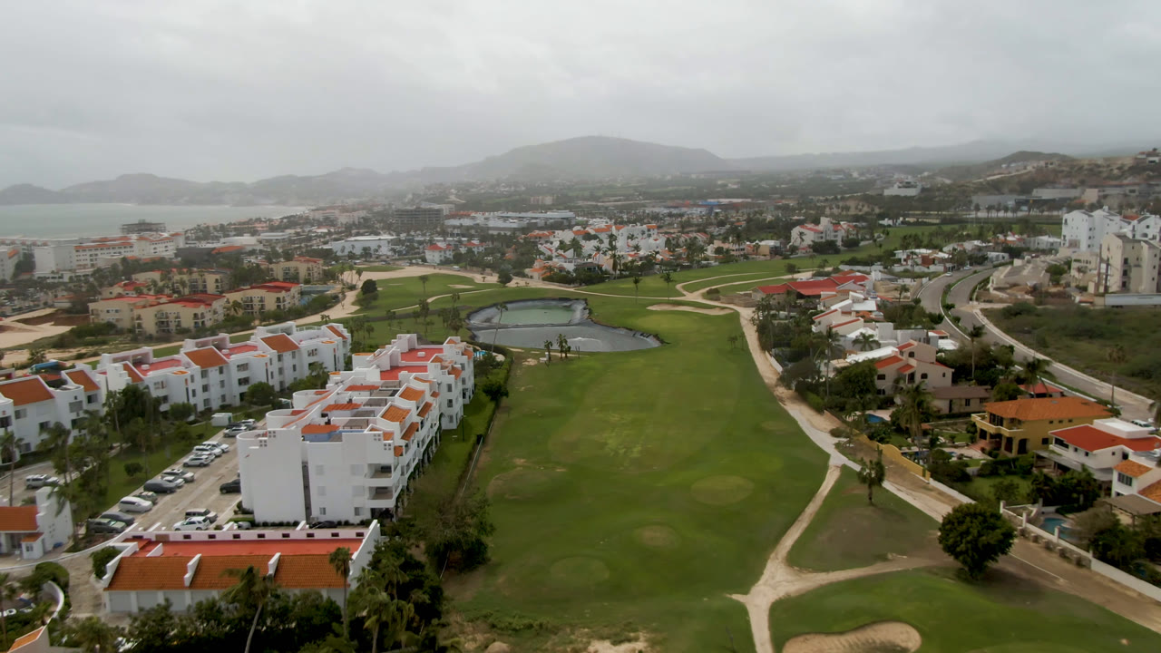 Opening aerial frame over a San Jose del Cabo golf course with white buildings at left, green fairways through the center, hazy hills and distant water beyond, and no visible people.