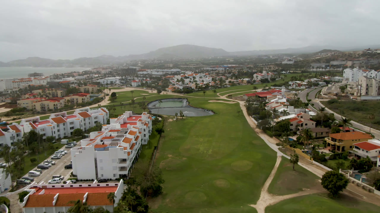 Aerial frame in San Jose del Cabo showing a central green fairway, a small pond left of center, white residential-style buildings, roads, hills, distant water, and no visible people.