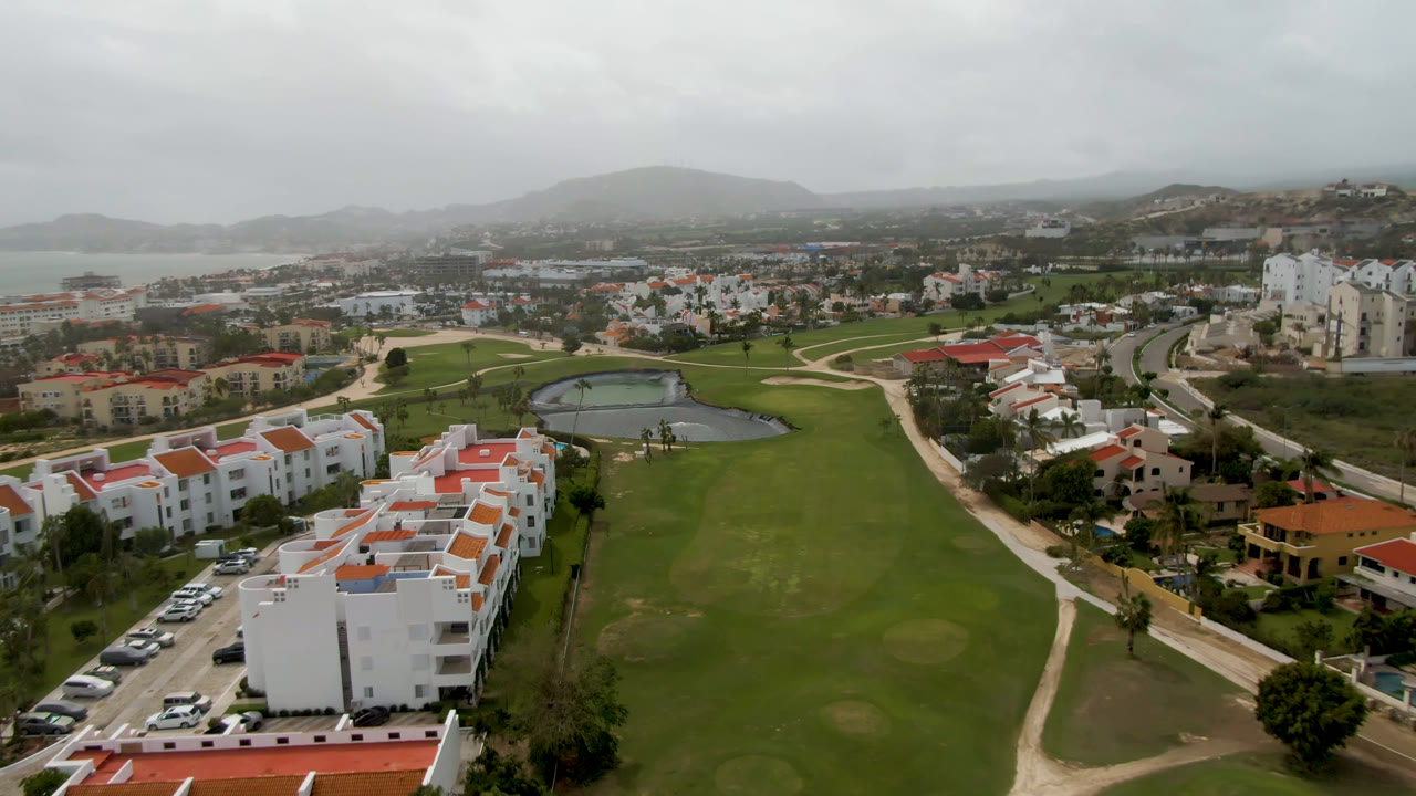Aerial golf course view with fairways, pond, white buildings, roads, and distant coastline.