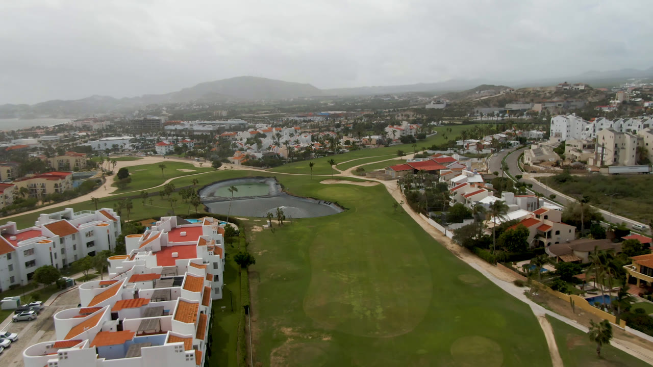 Wide aerial view of a golf course with a pond, white buildings, roads, and distant hills.