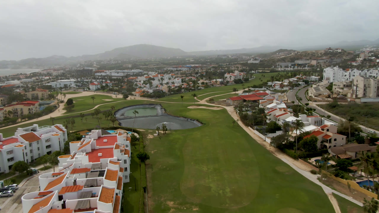 A second aerial frame shows the San Jose del Cabo fairways and pond from a similar high angle, with white buildings closer to the lower edge and no visible people.