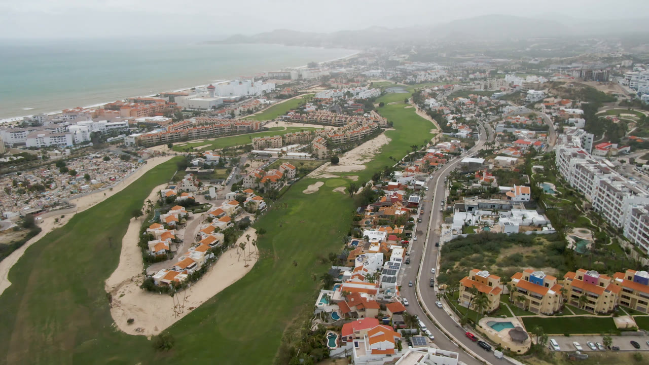 Opening aerial frame over San Jose Del Cabo shows green fairways, sandy bunkers, beachfront water, a curving road, low-rise buildings, hazy hills, and no visible people.