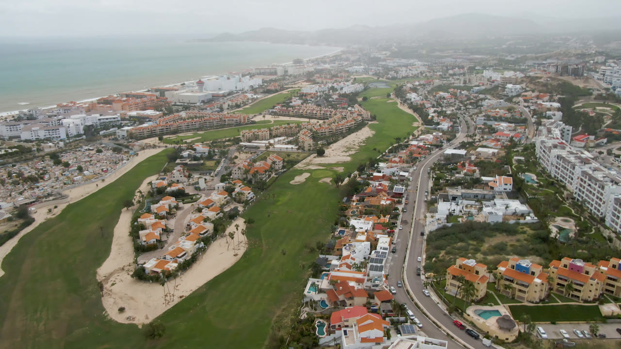 At 0:01, the San Jose Del Cabo aerial holds on fairways and sand traps in the foreground, white and red-roofed buildings along the road, ocean in the distance, and no visible people.