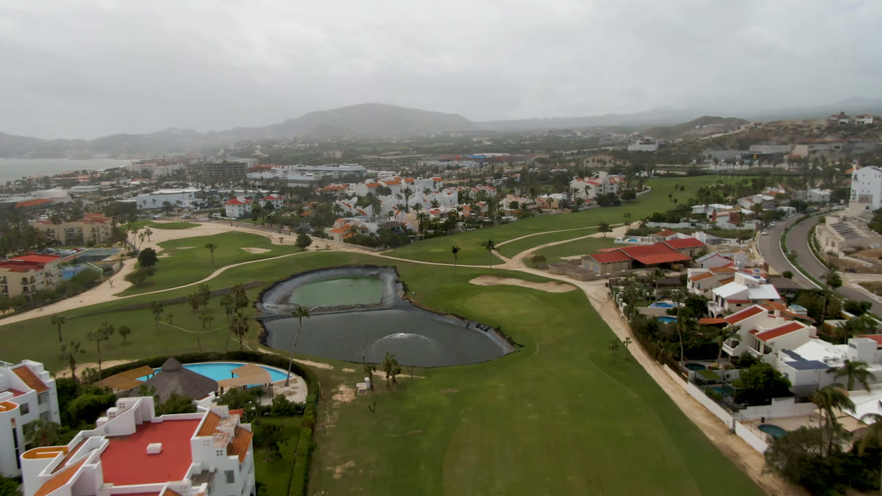 Aerial view of a golf course pond, fairways, pool, homes, roads, and distant hills under a hazy sky.