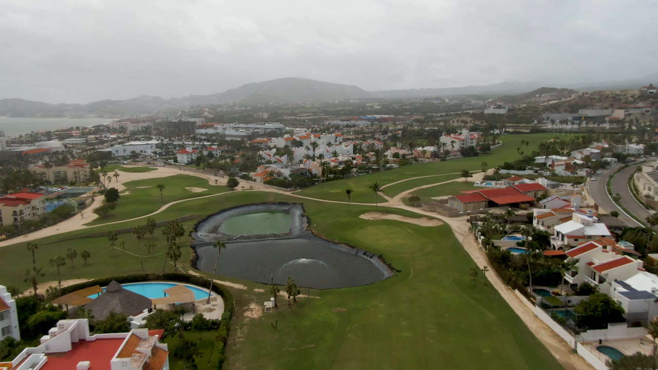 San Jose del Cabo aerial frame with the pond centered lower in view, fairways and bunkers spreading around it, residential buildings beside a road, and no visible people.
