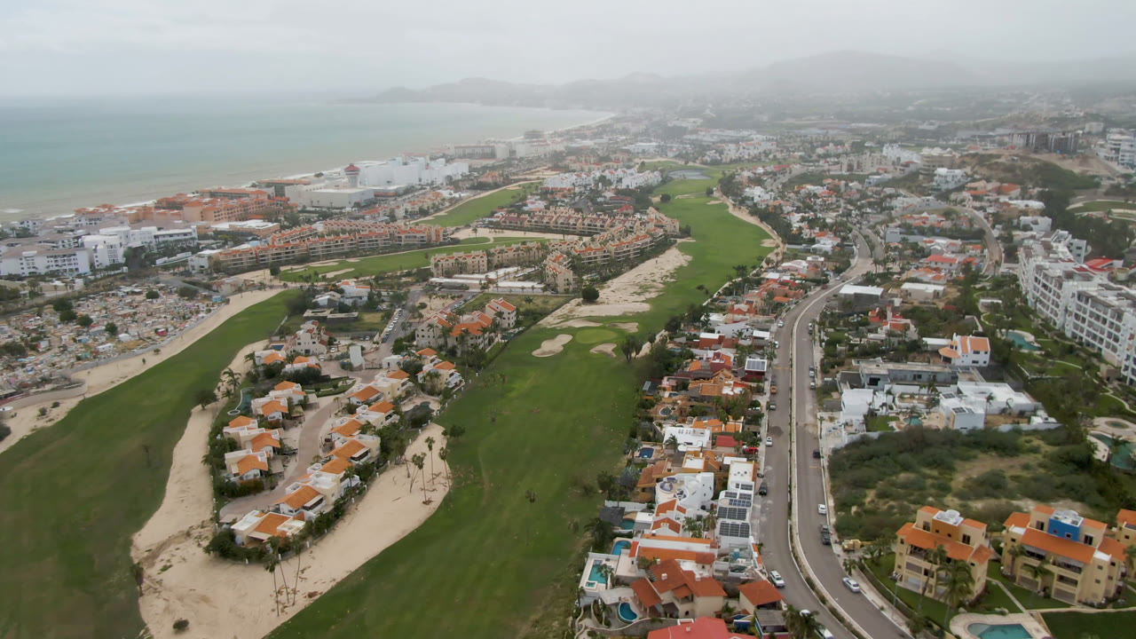 Aerial frame over San Jose del Cabo showing green golf fairways, sand traps, terracotta-roof homes, a coastal road, hazy ocean water, and no visible people.