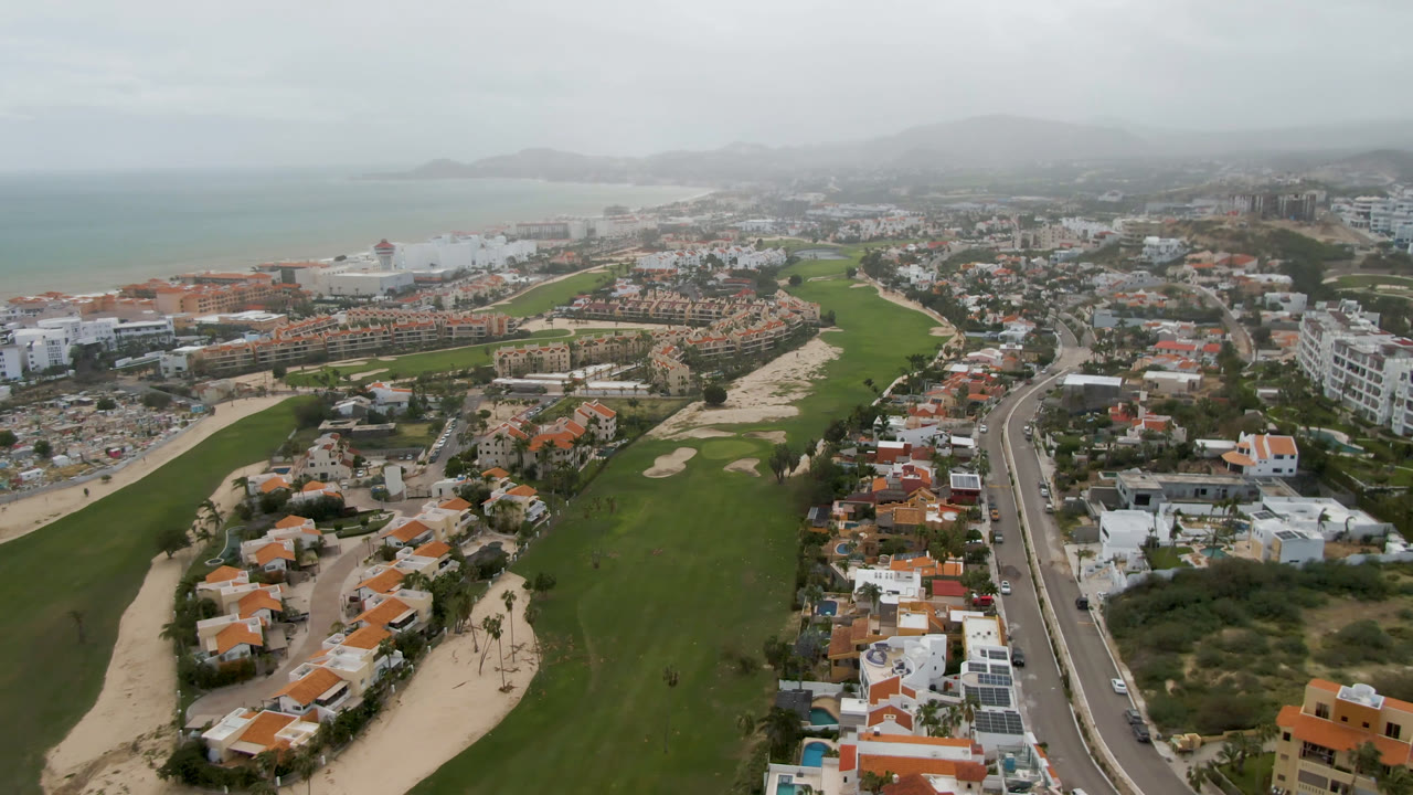 Aerial view of golf fairways, homes, road, coastline, and hazy hills.