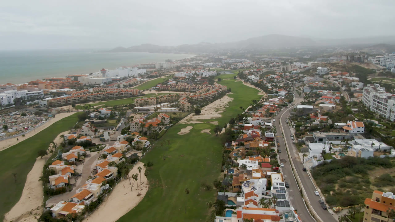 Second aerial frame in San Jose Del Cabo with the fairways centered, homes and roadside buildings along both sides, pale ocean in the background, and no visible people.