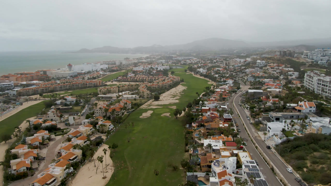 Opening aerial frame over San Jose del Cabo shows a green golf course, orange-roofed homes, a road, beach, pale sea, distant hills, and no visible people.