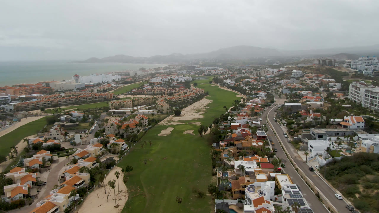 San Jose del Cabo aerial frame shows the fairway running toward the coast, with residential blocks, a curving road, beach, water, hazy hills, and no visible people.