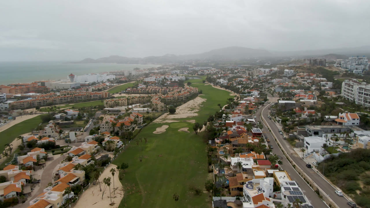 Wide aerial over golf course, houses, hotels, beach, sea, and overcast sky.