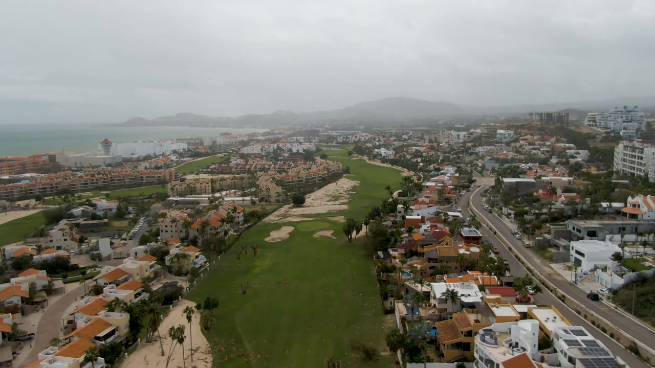 Aerial view of a golf course beside homes, roads, coastline, and hills under gray cloud.
