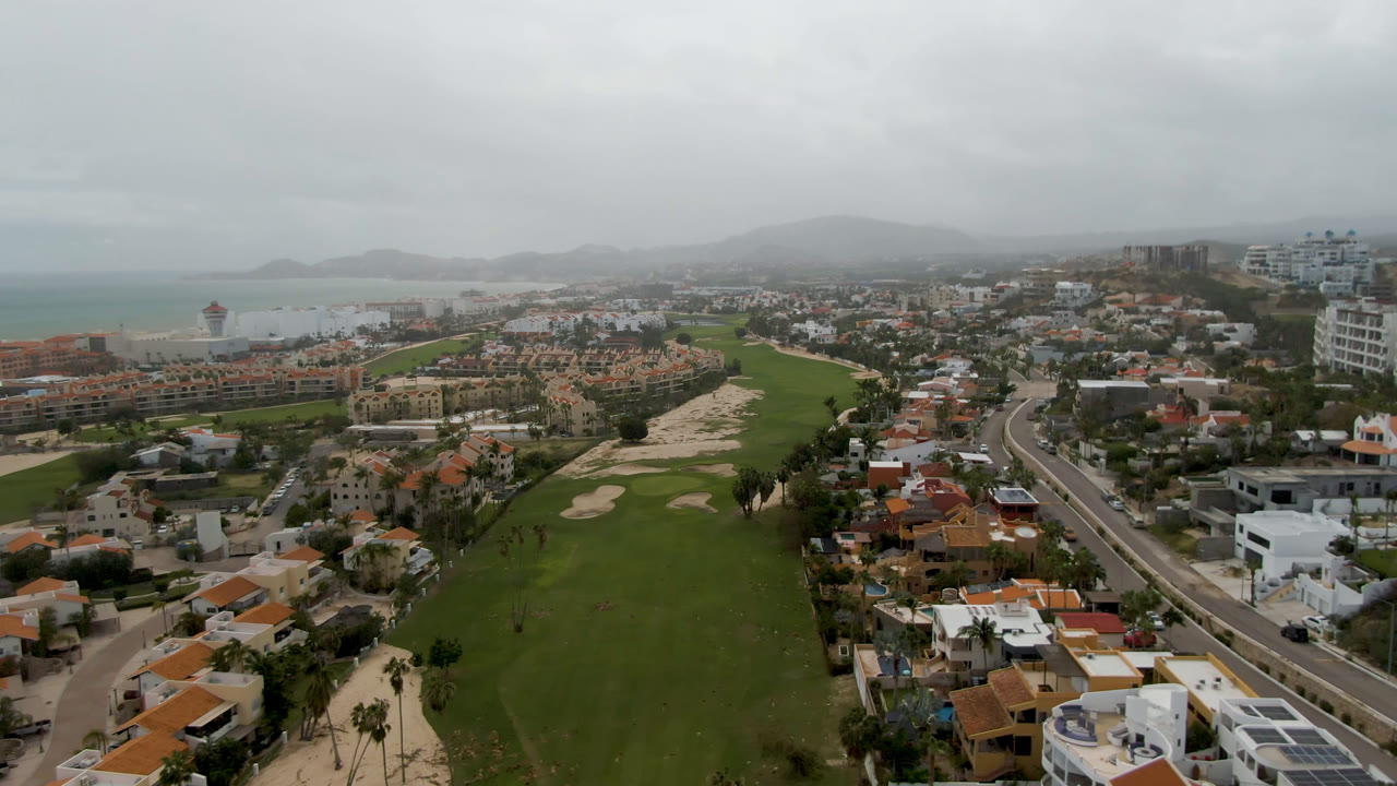 One second into the San Jose del Cabo aerial, the fairway fills the lower center while terracotta-roofed homes, a coastal strip, distant hills, and no visible people remain in view.