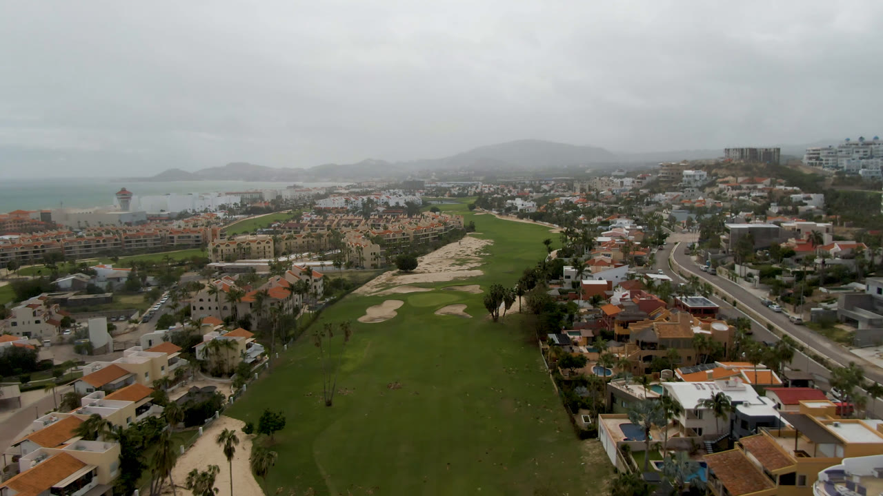 Wide aerial view of a golf course, homes, road, coastline, and cloudy hills.