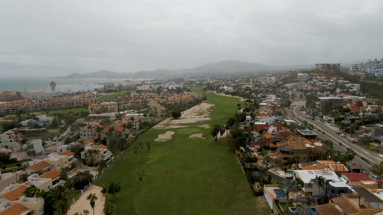 A second aerial frame in San Jose del Cabo holds on the green fairway, sand bunkers, road corridor, orange-roofed homes, distant coast, and no visible people.