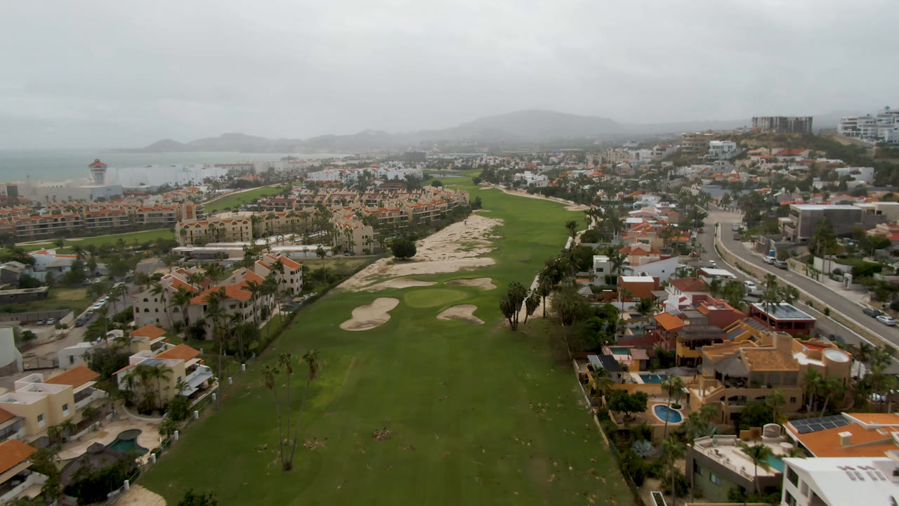 Aerial view over San Jose del Cabo with green golf fairways, sand bunkers, red-roofed buildings, distant ocean, hazy hills, gray clouds, and no visible people.