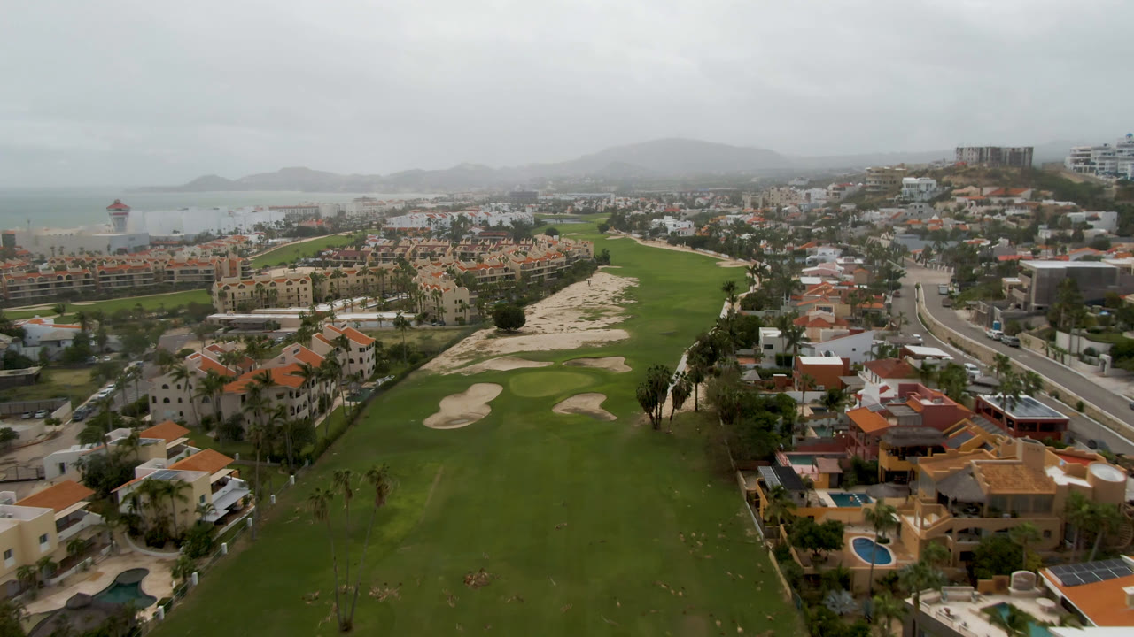 Aerial frame in San Jose del Cabo showing a broad green fairway, pale sand traps, villas with red roofs, a curving road, ocean in the distance, and no visible people.