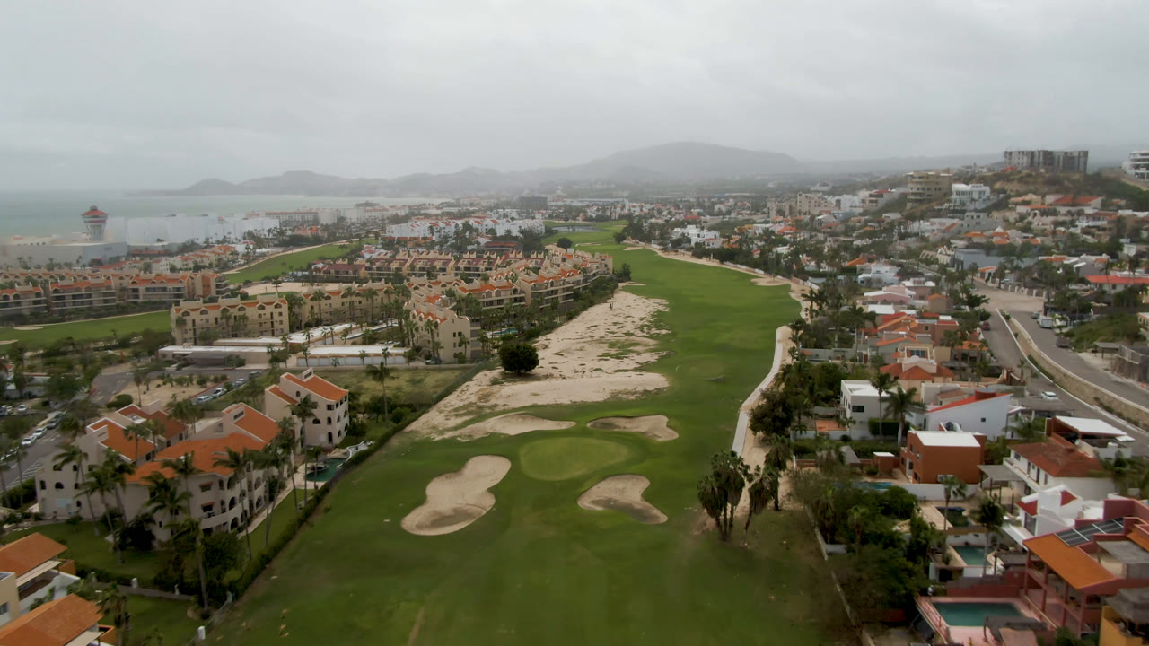 Opening aerial frame over a San Jose del Cabo golf course with green fairways, pale sand traps, orange-roofed villas, ocean water, distant hills, and no visible people.