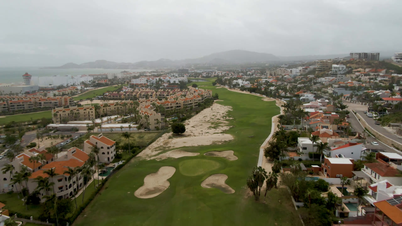Second aerial frame in San Jose del Cabo showing the fairway corridor, clustered sand bunkers, resort-style buildings, coastal water, hazy hills, and no visible people.