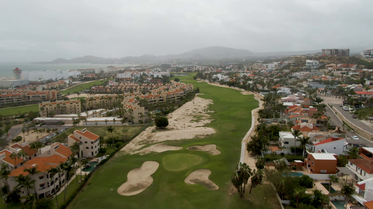 High aerial over a golf course bordered by villas, roads, ocean, and low hills.