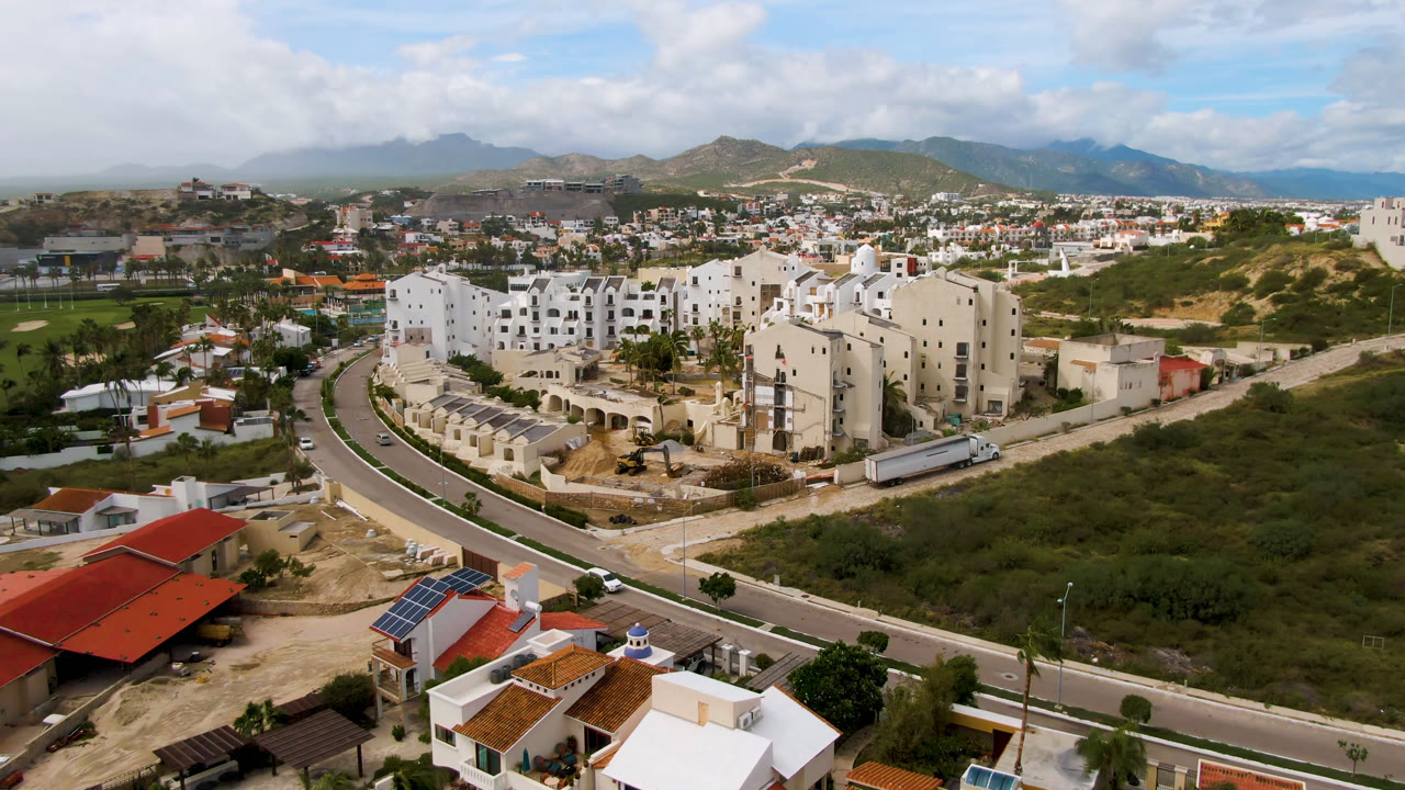 Opening aerial frame of San Jose del Cabo showing white apartment-style buildings near a curved road, red-roof homes in the foreground, low green terrain, distant hills, and no visible people.
