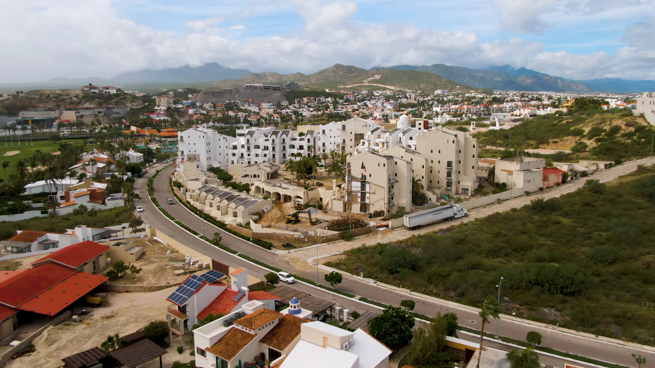 One second into the San Jose del Cabo aerial, the frame centers white residential buildings and roadside scrubland, with red and white rooftops below, hills beyond, and no visible people.