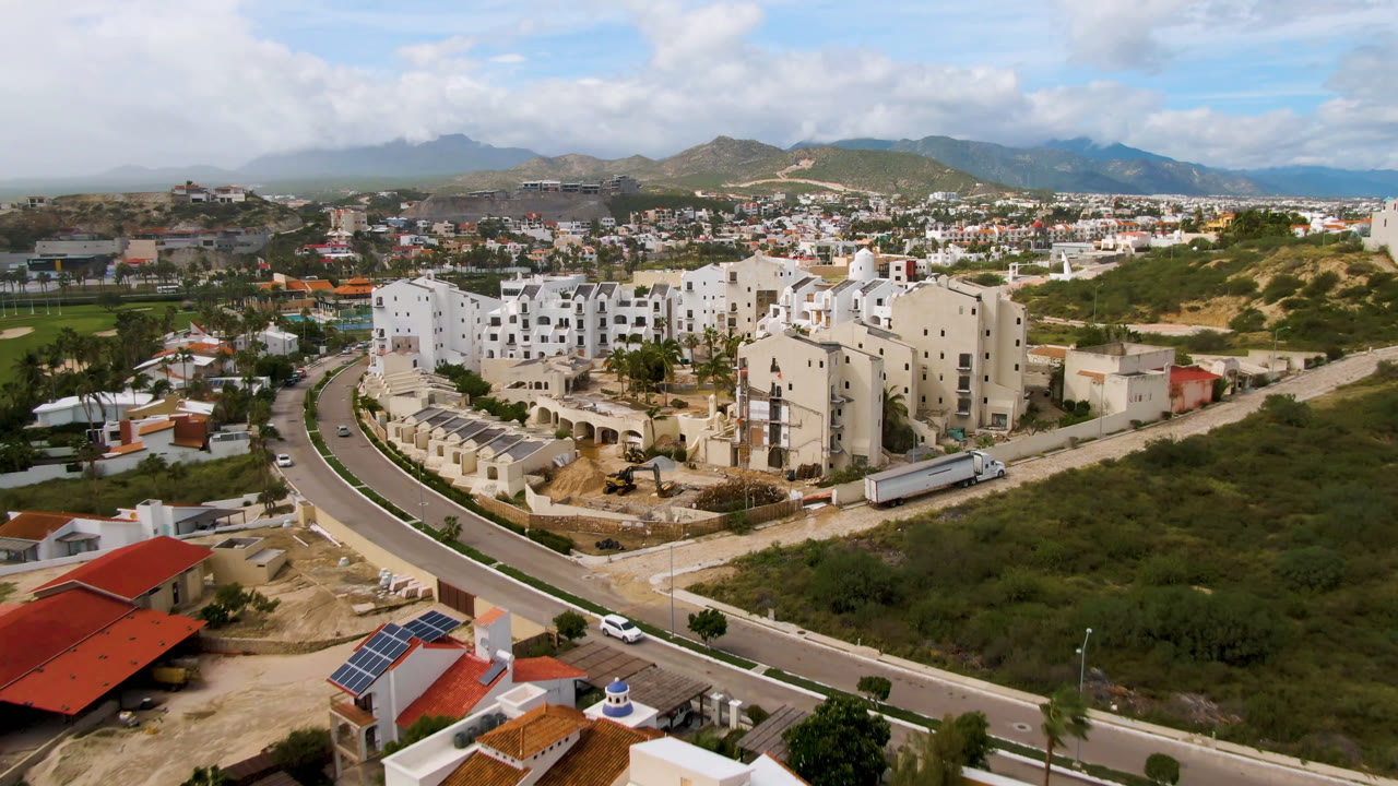 Aerial city view with white buildings, a curved roadway, small houses, scrubland, and mountains.