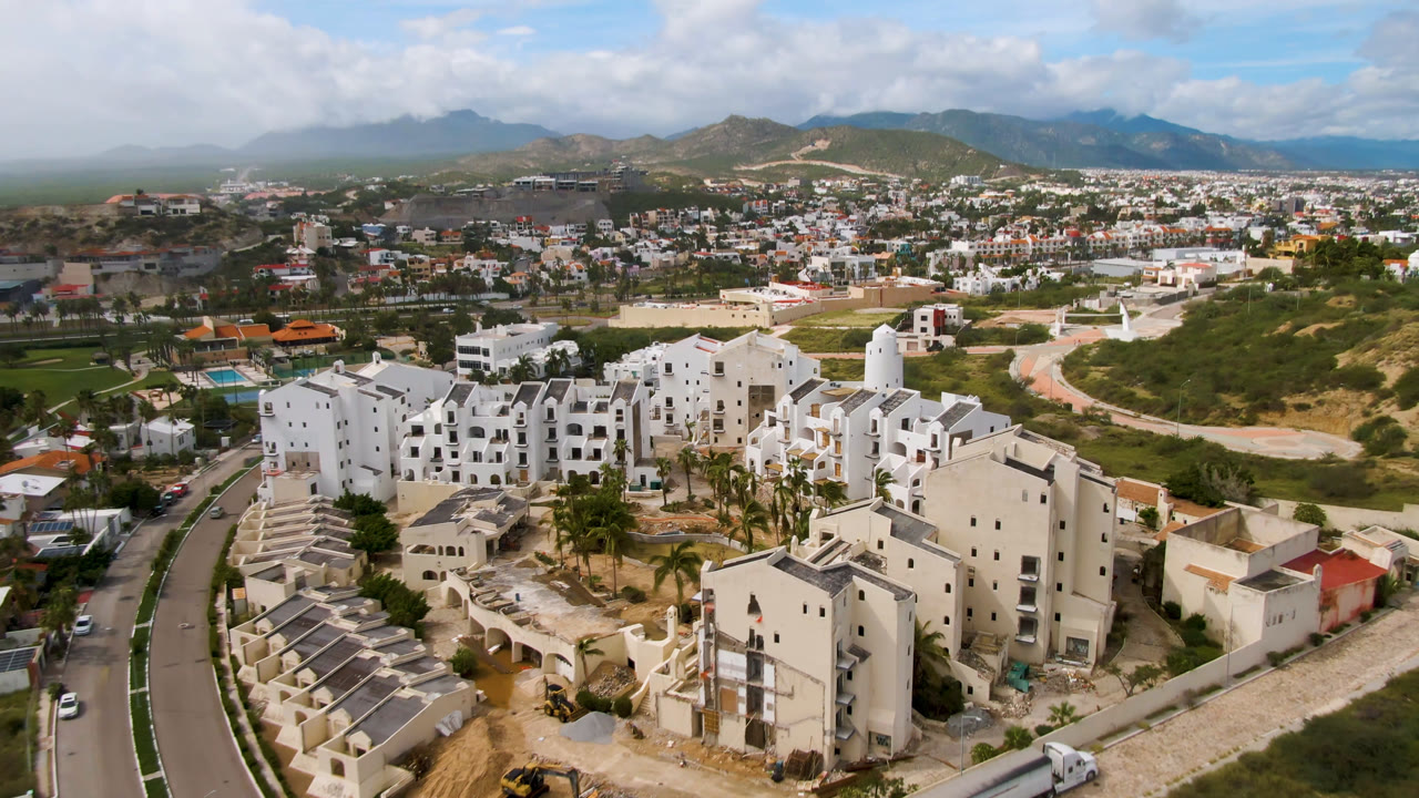 High aerial frame of San Jose del Cabo showing dense white buildings, a central road, scattered vehicles, green areas, mountain background, and no visible people.