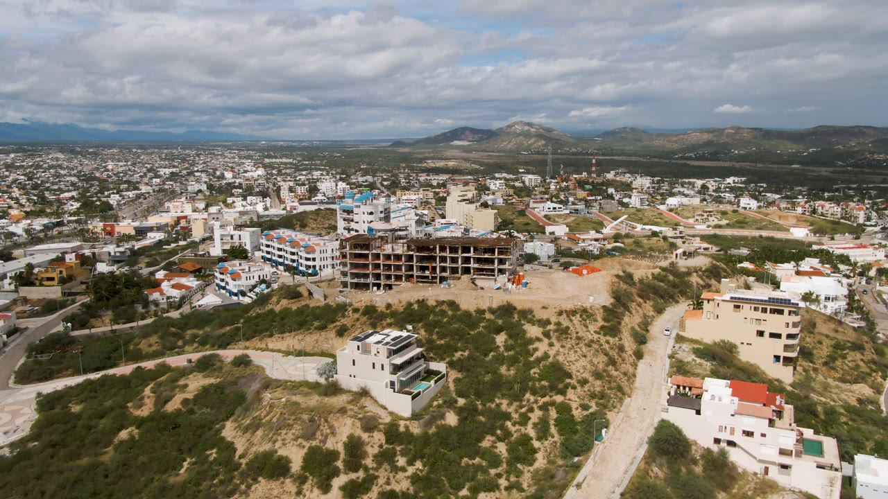 Wide aerial view of hillside homes, city blocks, construction, and distant mountains under clouds.