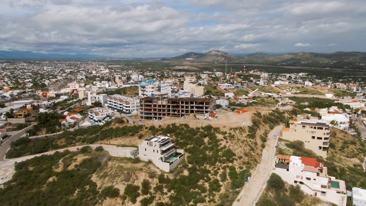 Second aerial frame over San Jose del Cabo, Mexico, with the construction structure near center, residential streets spreading behind it, hillside houses in front, and no visible people.