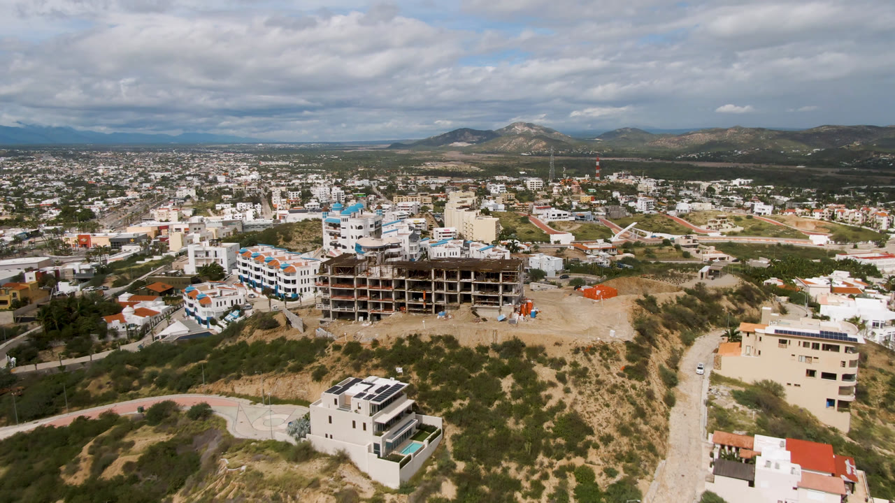 First frame of San Jose del Cabo from above, with a dry hillside in front, a large unfinished building near center, city blocks beyond, distant mountains, cloudy sky, and no visible people.