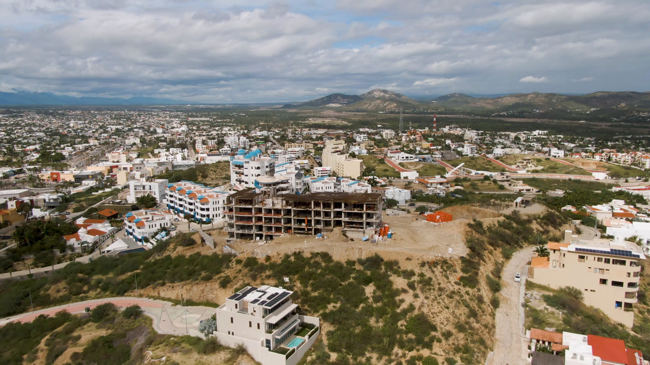 San Jose del Cabo aerial frame with the unfinished structure slightly more central, white and blue buildings to the left, scrubby slopes below, distant neighborhoods, mountains, and no visible people.