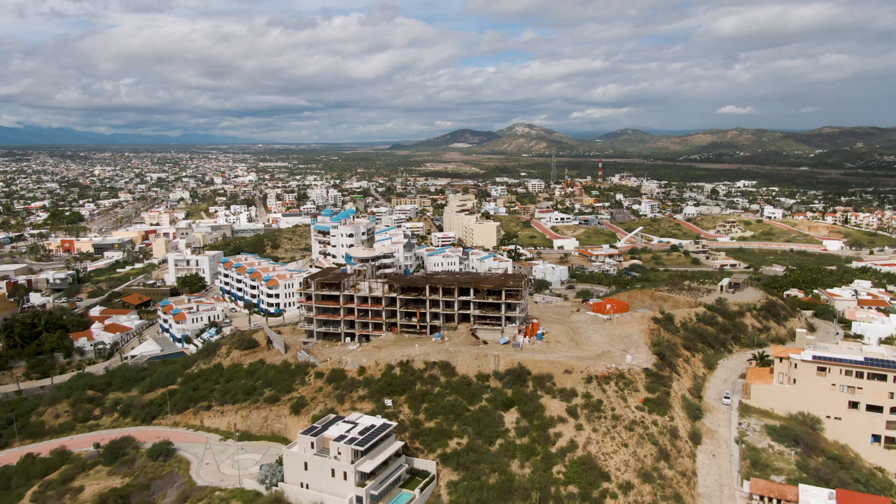 Wide aerial city view with an unfinished building, dry lots, roads, and mountains.