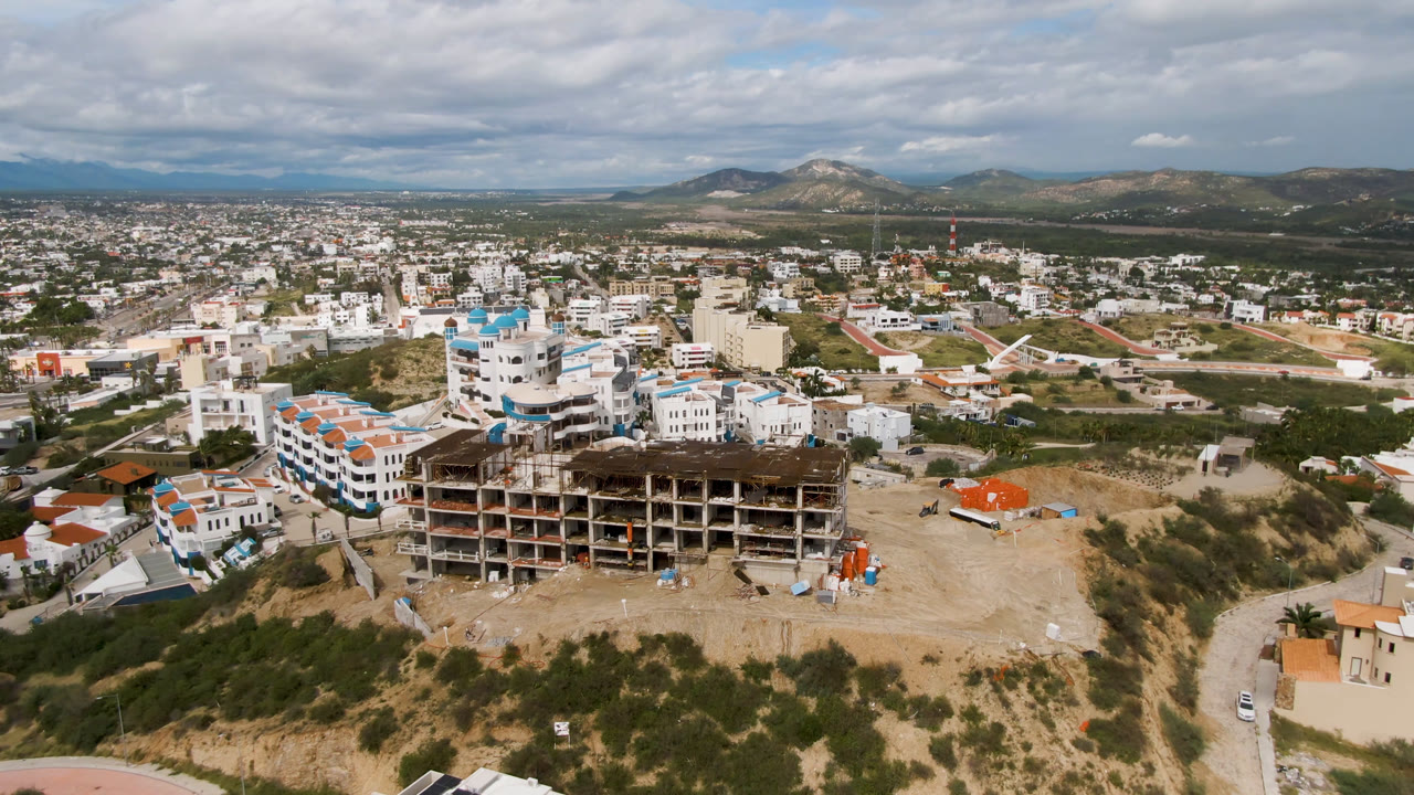 Aerial frame over San Jose del Cabo showing an unfinished concrete building on a dusty hillside, nearby white buildings, roads, scrub terrain, distant hills, and no visible people.