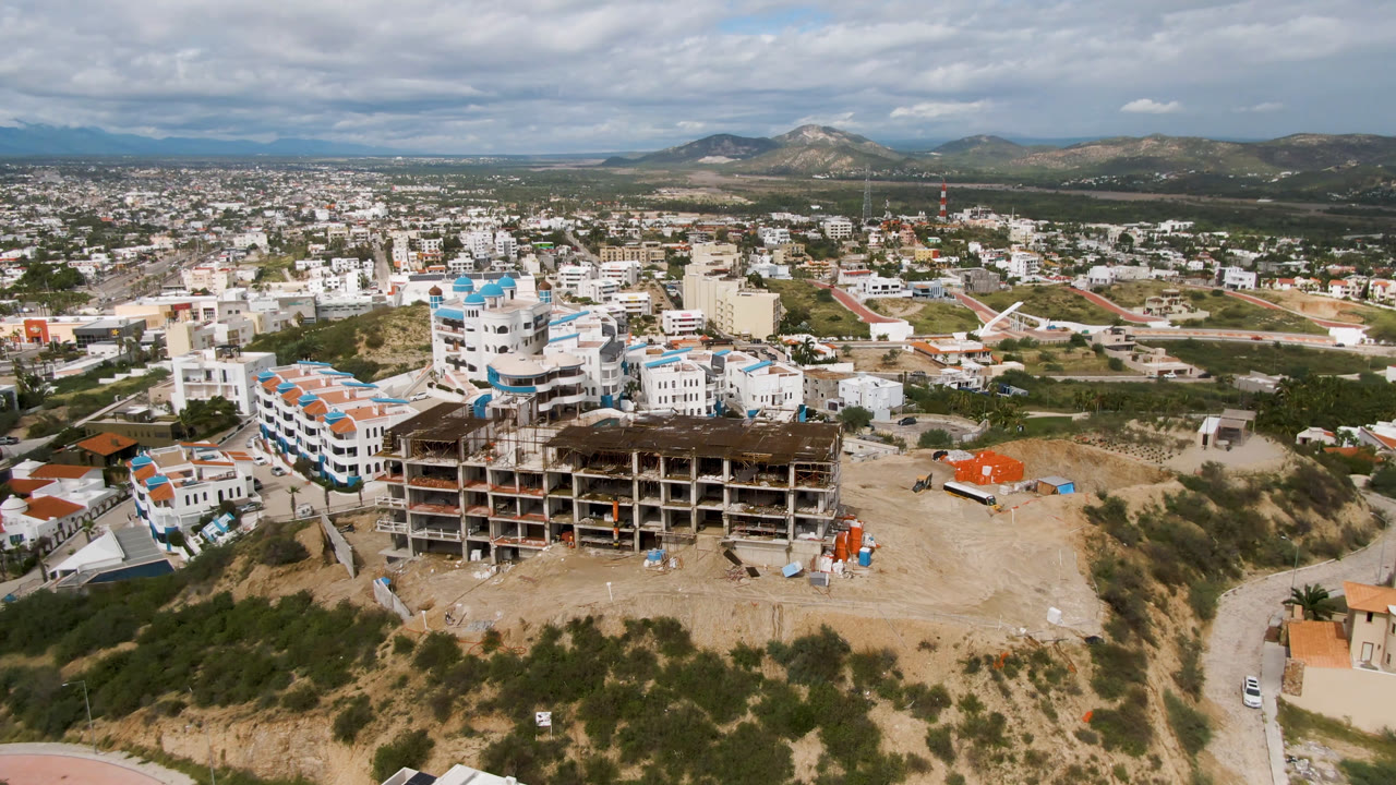 A second aerial view in San Jose del Cabo centers the skeletal construction structure, with resort-style white buildings to the left, exposed dirt lots, roads, mountains, and no visible people.