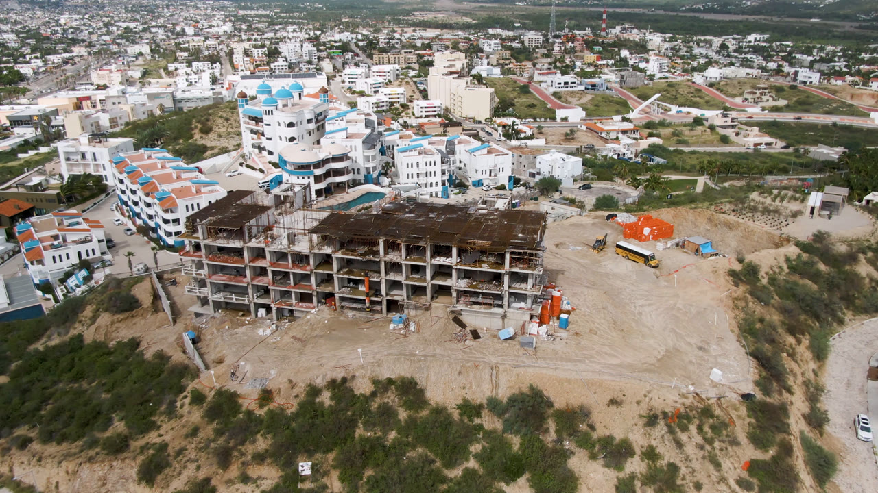 Aerial frame in San Jose Del Cabo showing a multi-level concrete construction site in the foreground, white buildings behind it, a yellow bus on a dirt road, and no visible people.