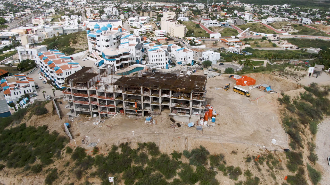 Aerial frame in San Jose Del Cabo with the unfinished concrete structure centered lower in frame, red-roofed white buildings to the left, orange-covered materials to the right, and no visible people.