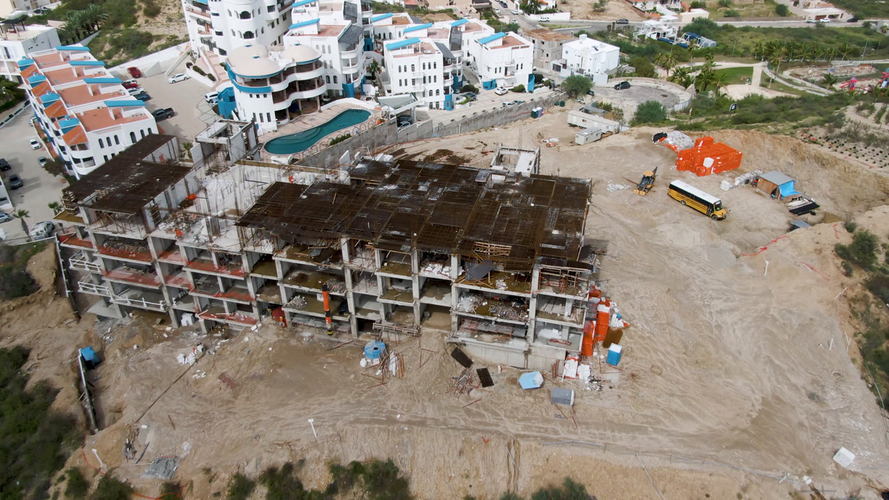 Aerial frame in San Jose del Cabo looking over the unfinished building, cleared dirt lot, yellow bus, orange construction materials, adjacent roads, white buildings, and no visible people.
