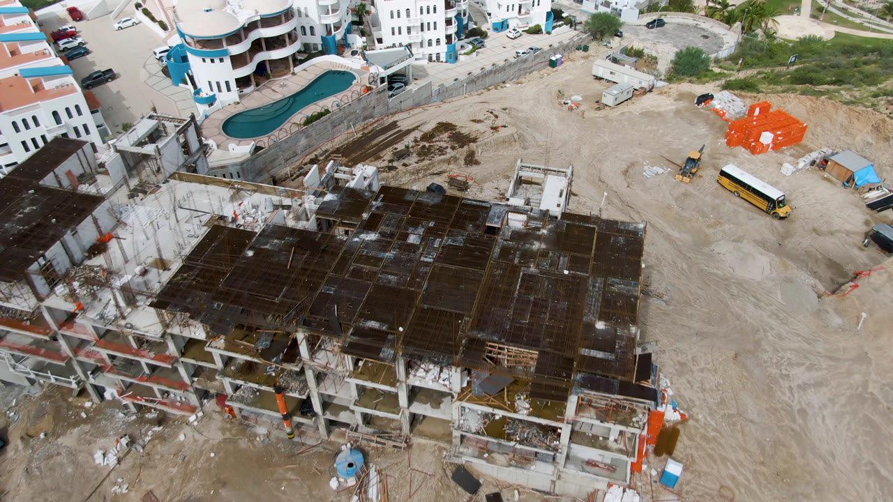 Overhead view of a San Jose del Cabo construction site with a dark concrete roof in the foreground, a dirt lot, a curved pool, orange material stacks, parked vehicles, and no visible people.