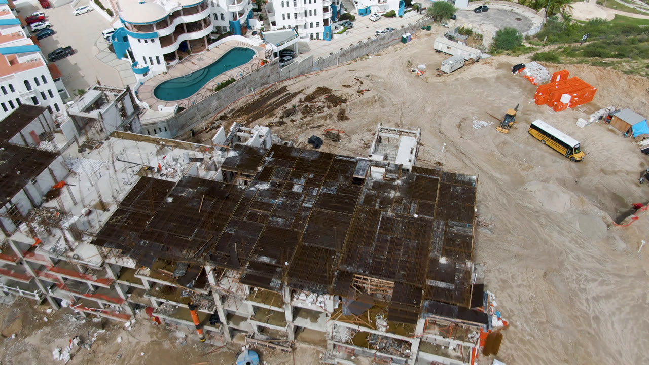 High-angle frame in San Jose del Cabo showing the unfinished building deck across the lower half, open graded ground, a curved pool near white buildings, a yellow bus, and no visible people.