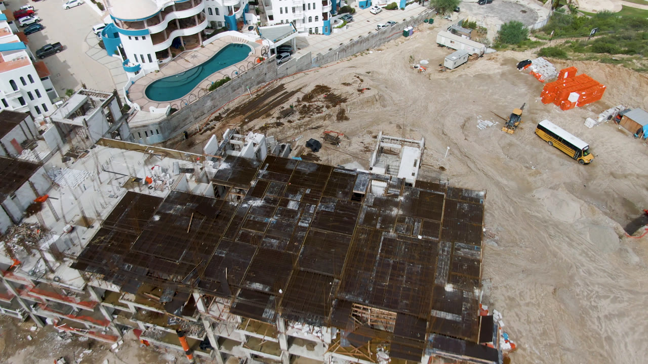Aerial construction site beside white buildings, pool, vehicles, and material stacks.